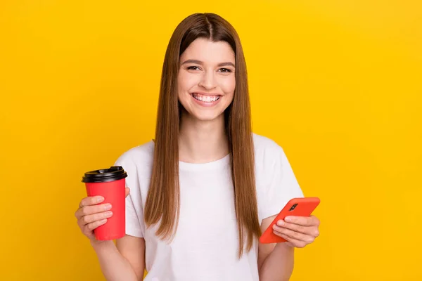 Фото молодой счастливой позитивной женщины улыбка держать телефон вынос кофе улыбка изолированы на светло-желтый цвет фона — стоковое фото