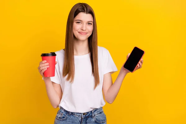 Фото позитивной молодой счастливой женщины улыбка показать телефон экран пустое пространство провести кофе изолированы на желтый цвет фона — стоковое фото