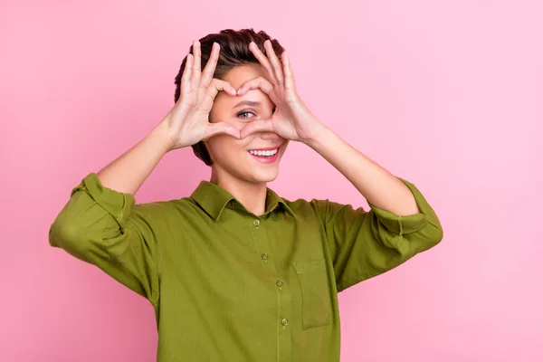 Bild av flirtig lekfull dam fingrar täcka ögat visa hjärta symbol bära grön skjorta isolerad rosa färg bakgrund — Stockfoto
