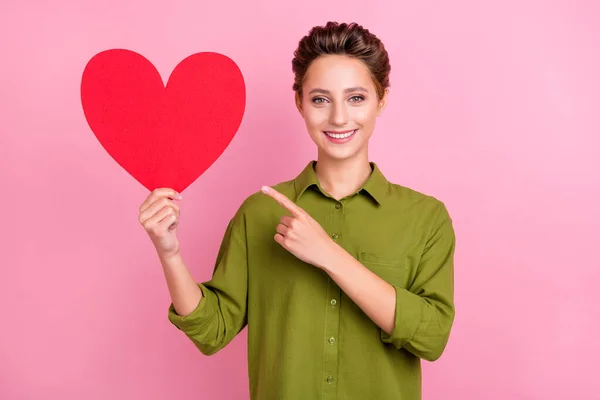 Фото милой блестящей молодой женщины, одетой в зеленую рубашку, улыбающейся указательным пальцем красное сердце пустое пространство изолированный розовый цвет фона — стоковое фото