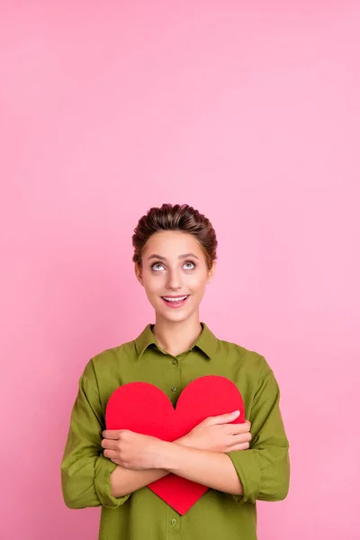 Photo verticale de mignonne impressionnée jeune femme habillée chemise verte étreignant coeur rouge regardant espace vide isolé fond de couleur rose — Photo
