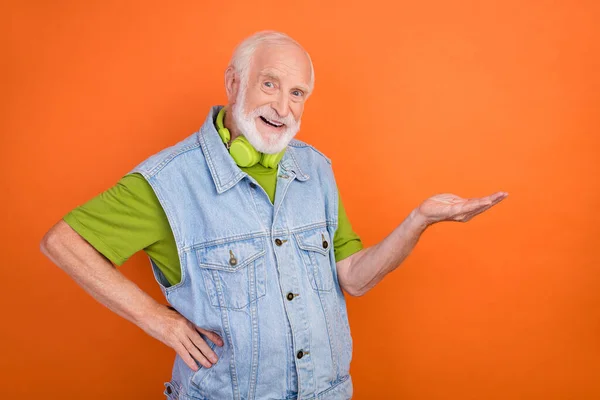 Photo of charming sweet man pensioner dressed retro denim vest earphones smiling holding arm empty space isolated orange color background — Stock Photo, Image