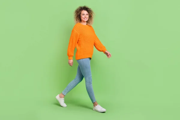 完全な長さ写真のかわいいブロンド波毛の女性行く服オレンジプルオーバージーンズ履物隔離された緑の背景 — ストック写真