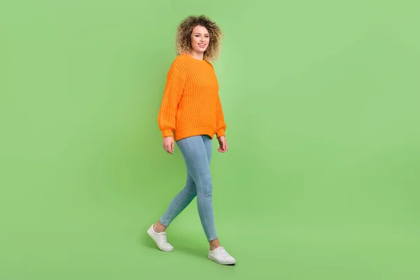 フルサイズ写真の素敵なブロンド巻き毛の女性行く服オレンジセータージーンズ履物隔離された緑の背景 — ストック写真