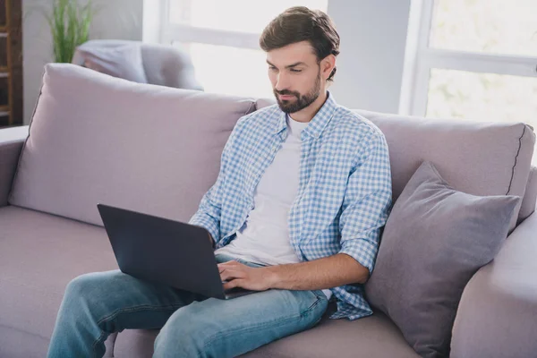 Foto van positieve baardgerichte freelancer man zitten bank typen pc e-mail dragen casual outfit in comfortabel huis binnen — Stockfoto