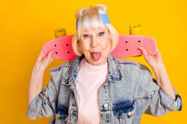 Фотопортрет пожилой женщины, держащей скейтборд, показывающий язык, чувствует себя молодым изолированным на ярко-желтом фоне — стоковое фото