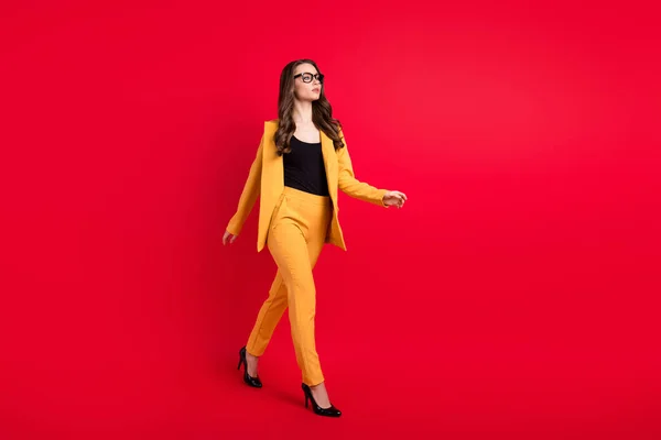 Foto de mujer joven seria brillante usar gafas chaqueta amarilla caminando espacio vacío aislado color rojo fondo —  Fotos de Stock