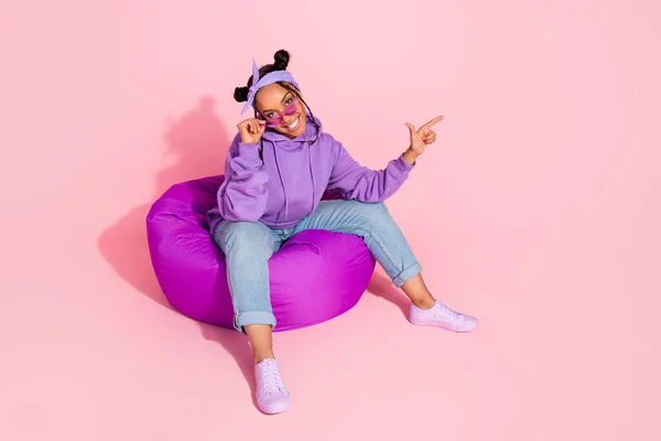 Foto de dulce dama africana fresca vestida violeta sudadera bollos brazo gafas frijol bolso apuntando espacio vacío aislado color rosa fondo —  Fotos de Stock