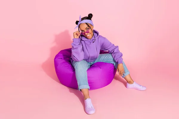 Foto de mulher afro atraente funky usar roxo pães com capuz sentado beanbag braço óculos sorrindo isolado cor de fundo rosa — Fotografia de Stock