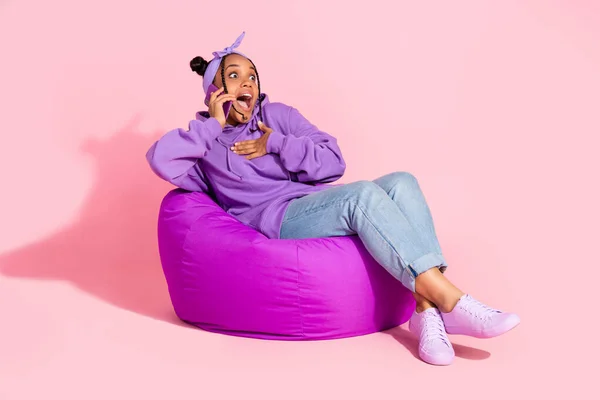 Foto de atractiva hermosa dama africana vestida violeta sudadera bollos hablando moderno gadget sentado frijol bolso aislado de color rosa fondo —  Fotos de Stock