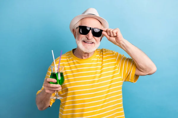 Komik yaşlı beyefendinin fotoğrafı sarı t-shirt şapkalı koyu gözlükler takıyor. Kokteyl tutarken gülümsüyor. Mavi arka plan. — Stok fotoğraf