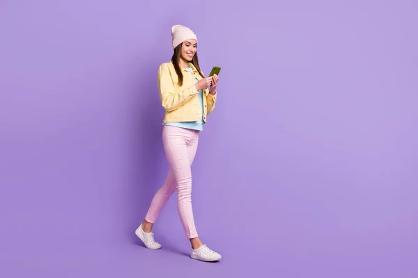 Full storlek profil foto av optimistiska raka frisyr flicka gå titta telefon bära mössa jacka byxor sneakers isolerade på lila bakgrund — Stockfoto