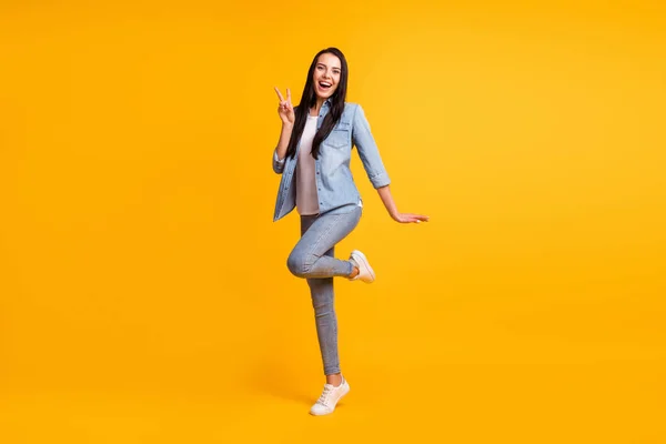 Full size foto van mooie optimistische brunette dame show v-teken dragen blauwe shirt jeans sneakers geïsoleerd op gele kleur achtergrond — Stockfoto