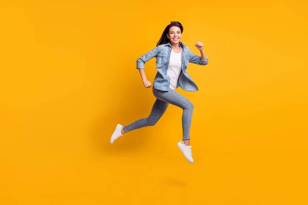 Full size profiel foto van cool optimistische brunette dame run springen dragen blauw shirt jeans sneakers geïsoleerd op gele achtergrond — Stockfoto