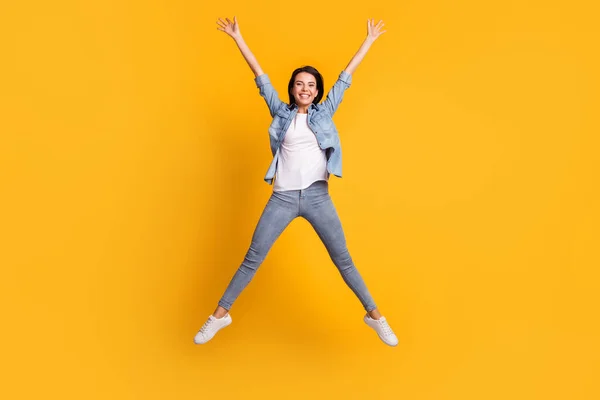 Foto de tamaño completo de agradable optimista dama morena salto usar pantalones vaqueros camisa azul zapatillas aisladas sobre fondo amarillo —  Fotos de Stock