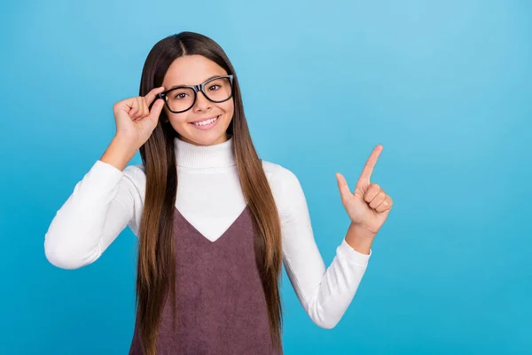 Foto av roliga smarta preteen flicka klädd brun övergripande arm glasögon pekar tomt utrymme leende isolerad blå färg bakgrund — Stockfoto