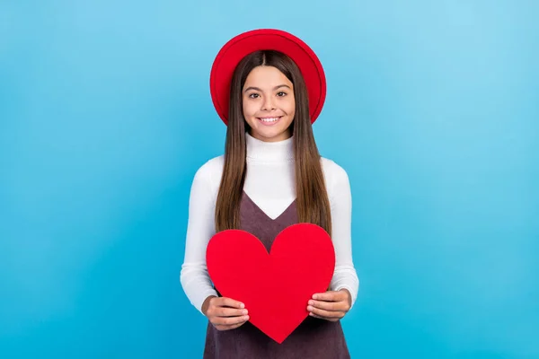 Foto de doce bonito preteen menina vestida marrom geral vermelho headwear segurando grande coração sorrindo isolado azul cor de fundo — Fotografia de Stock