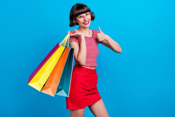 Potret gadis tersenyum menjaga tas setelah berbelanja menunjukkan ibu jari-up terisolasi warna biru latar belakang — Stok Foto
