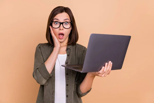 Photo of crazy shocked lady hold laptop dłoń policzek otwarte usta nosić okulary khaki koszula odizolowany beżowy kolor tło — Zdjęcie stockowe