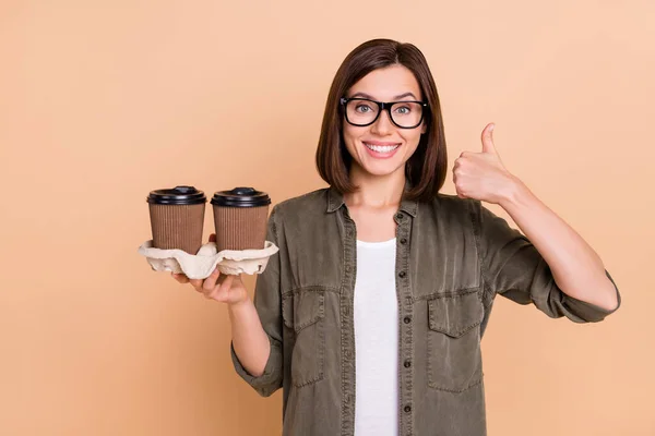 Foto de garçonete senhora segurar takeout café levantar polegar até especificações de desgaste camisa cáqui isolado cor bege fundo — Fotografia de Stock