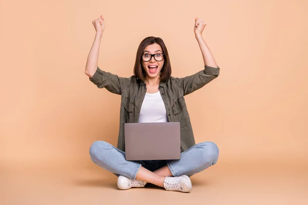 Фото леді сидить підлога тримає ноутбук радісні підняті кулаки носити окуляри хакі сорочки ізольовані бежевий колір фону — стокове фото