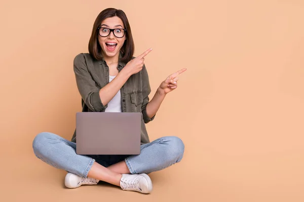 Photo of funny lady sit floor hold pc direct finger empty space open mouth wear khaki shirt isolated beżowy kolor tło — Zdjęcie stockowe