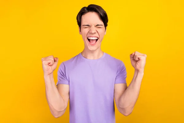 Foto de chico campeón asombrado levantar los puños cerrar los ojos gritar usar púrpura camiseta aislado color amarillo fondo — Foto de Stock