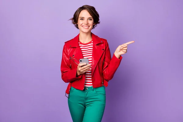 Fénykép a fiatal lány boldog mosoly használata mobiltelefon jelzi ujj üres tér tanácsot választani hirdetés elszigetelt felett lila színű háttér — Stock Fotó