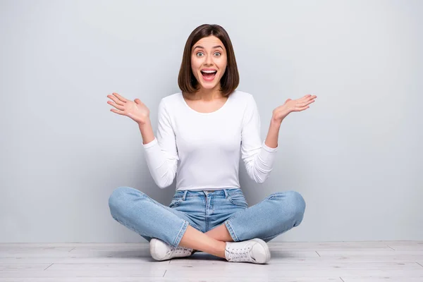 Foto von niedlichen beeindruckt junge Frau tragen weißes Outfit steigende Arme sitzen Beine gekreuzt lächelnd isoliert Beton grau Wand Hintergrund — Stockfoto