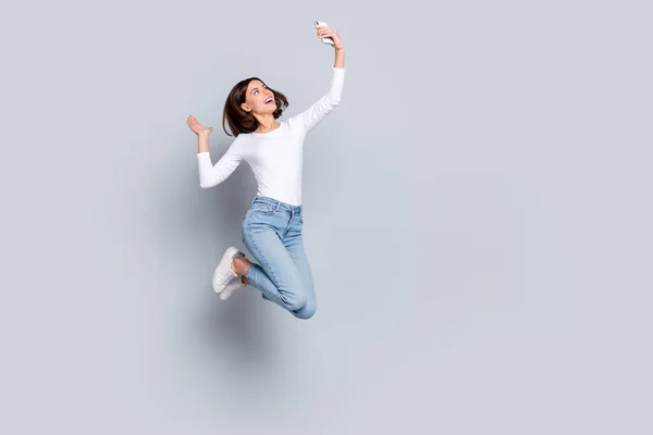 Parlak, şirin bir kadının fotoğrafı beyaz kıyafetler giyer yüksek atlama selfie modern cihaz boş uzay gülümsemesi izole edilmiş gri arka plan. — Stok fotoğraf