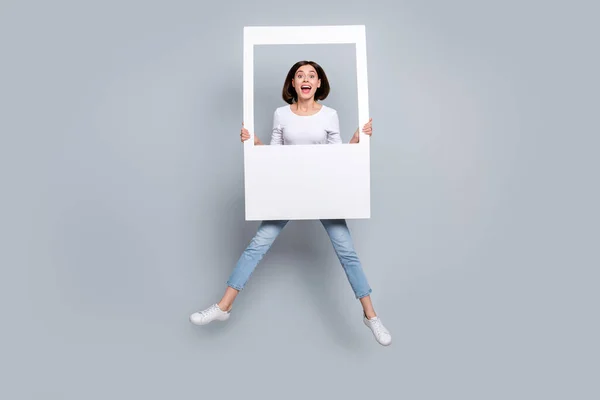 Foto de doce impressionado jovem mulher usar roupa branca saltando alta segurando moldura branca foto sorrindo isolado fundo de cor cinza — Fotografia de Stock