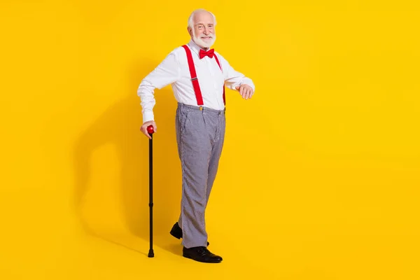 Teljes hosszúságú fotó vicces öreg ember bottal megy viselni nyakkendő ing nadrágtartó kockás nadrág cipő elszigetelt sárga színű háttér — Stock Fotó