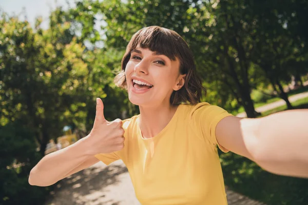 Foto de dulce joven excitada vestida camiseta amarilla caminando virando selfie mostrando el pulgar hacia arriba sonriendo al aire libre parque urbano — Foto de Stock