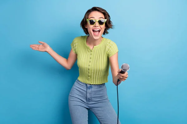 Foto de dulce mujer joven divertida vestida camiseta verde gafas oscuras invitando a cantar karaoke sonriendo aislado color azul fondo —  Fotos de Stock