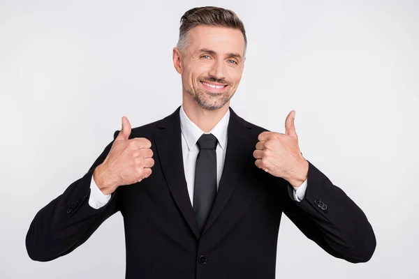 Фото позитивного жизнерадостного молодого менеджера в черной куртке, улыбающегося, показывающего большие пальцы на изолированном сером фоне — стоковое фото