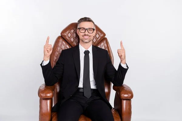 Foto van doordachte positieve jonge ondernemer dragen zwarte jas zitten lederen fauteuil wijzen lege ruimte geïsoleerde grijze kleur achtergrond — Stockfoto