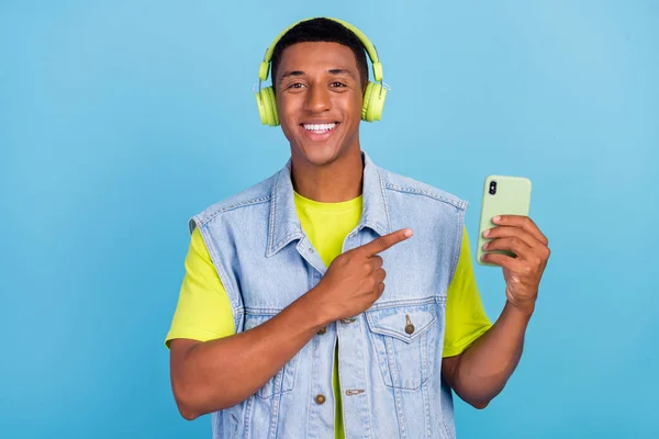Foto de joven afro hombre indicar dedo smartphone anuncios aconsejan auriculares aislados sobre fondo de color azul — Foto de Stock