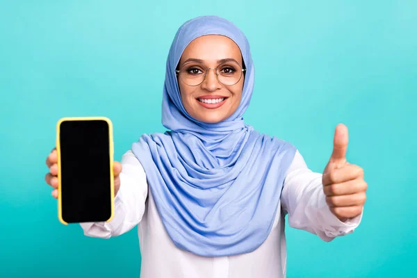 Foto di dolce brillante giovane signora vestita occhiali hijab islamici sorridente tenendo dispositivo moderno spazio vuoto pollice su isolato colore verde acqua sfondo — Foto Stock