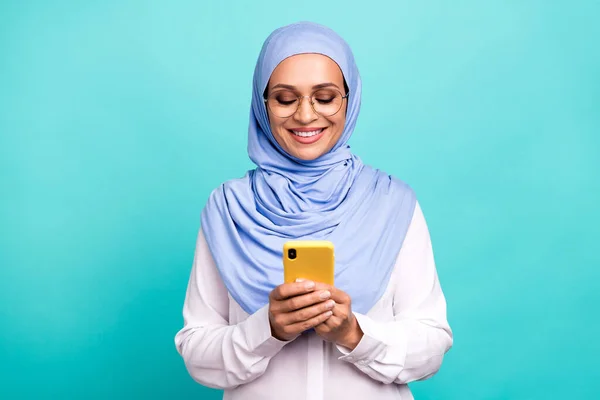 Fénykép fényes édes fiatal hölgy öltözött iszlám hidzsáb szemüveg mosolygós üzenetküldés modern eszköz elszigetelt varangy szín háttér — Stock Fotó
