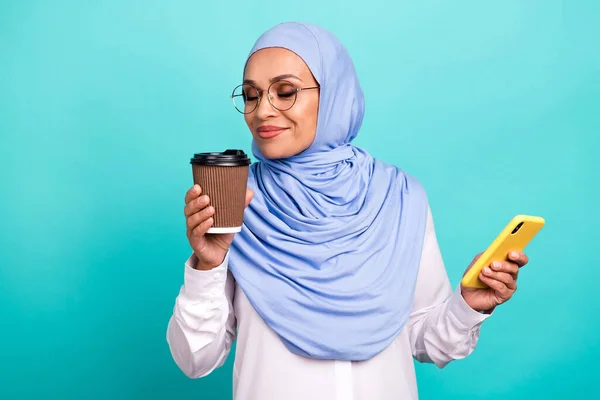 Foto de bonito muito jovem mulher desgaste árabe óculos de lenço de cabeça desfrutando de aroma de chá moderno gadget isolado cor turquesa fundo — Fotografia de Stock