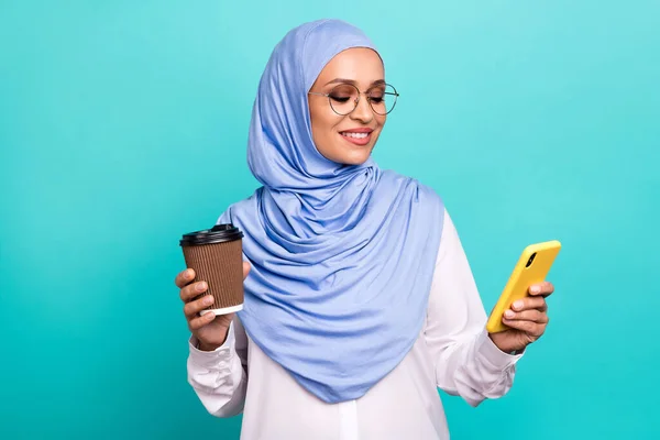 魅力的な光沢のある若い女性の写真を身に着けているイスラムヒジャーブ眼鏡笑顔タイプ現代的なデバイスを飲むカカオ孤立したティール色の背景 — ストック写真
