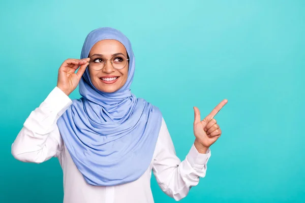 Foto de linda jovem senhora vestida islâmico hijab braço óculos sorrindo olhando apontando espaço vazio isolado cor teal fundo — Fotografia de Stock