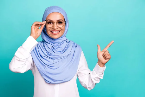 思慮深い自信のある若い女性の写真は、空のスペースを笑顔孤立したターコイズ色の背景を指すアラブのスカーフ腕の眼鏡を着用 — ストック写真
