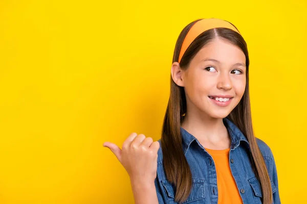 Foto de menina estudante sonhador funky usar camisa jeans sorrindo olhando apontando polegar para trás espaço vazio isolado cor amarela fundo — Fotografia de Stock