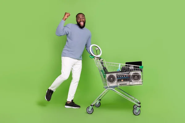 Fotografie šťastného africký chlap oblečený šedý svetr nákup moderní gadgets rostoucí pěst izolované zelené barvy pozadí — Stock fotografie