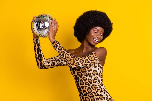 Фото возбуждённой темнокожей женщины, одетой в костюм леопарда, улыбающейся танцующей, держа диско-шар на изолированном жёлтом фоне — стоковое фото