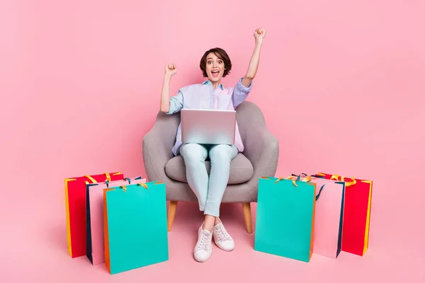 Longitud completa cuerpo tamaño foto mujer sentado con paquetes de gestos como ganador con portátil aislado pastel de color rosa de fondo —  Fotos de Stock
