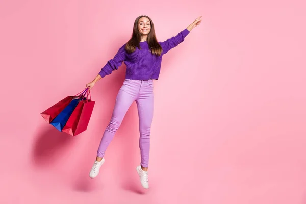 Фотографія повного розміру весела мила леді, що стрибає, тримає сумки прямим пальцем порожній простір ізольовано на рожевому кольоровому фоні — стокове фото