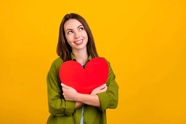 Foto de doce adorável jovem senhora usar camisa verde abraçando grande coração olhando espaço vazio sorrindo isolado cor amarela fundo — Fotografia de Stock
