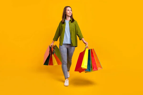 Fénykép a bájos fényes hölgy viselni zöld ing mosolygós séta gazdaság bevásárló táskák keres üres tér elszigetelt sárga háttér — Stock Fotó